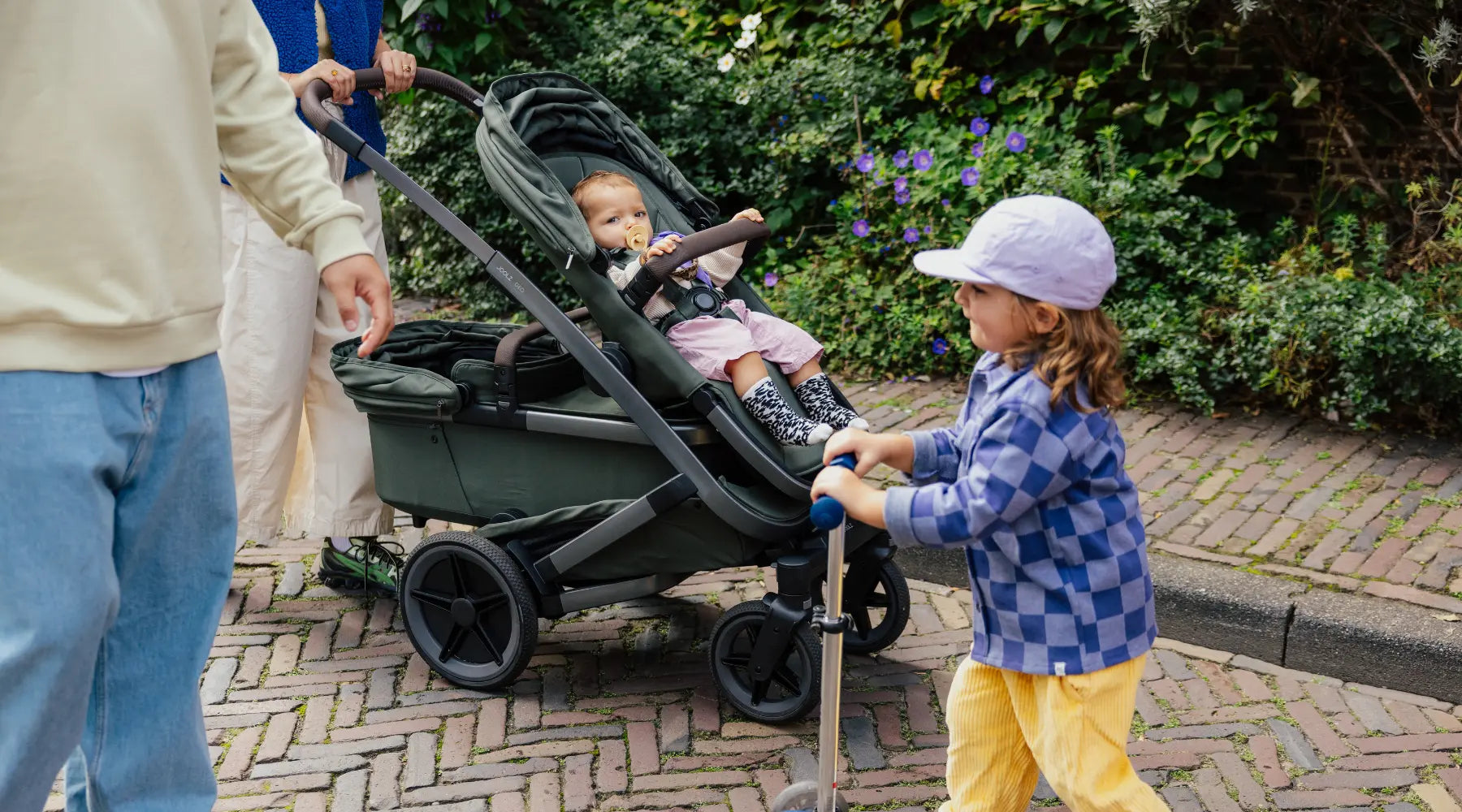 Eine Familie unterwegs: Der Joolz Geo5 Single in Forest Green mit Baby im Sportsitz auf Pflastersteinen, begleitet von einem Geschwisterkind auf einem Roller.