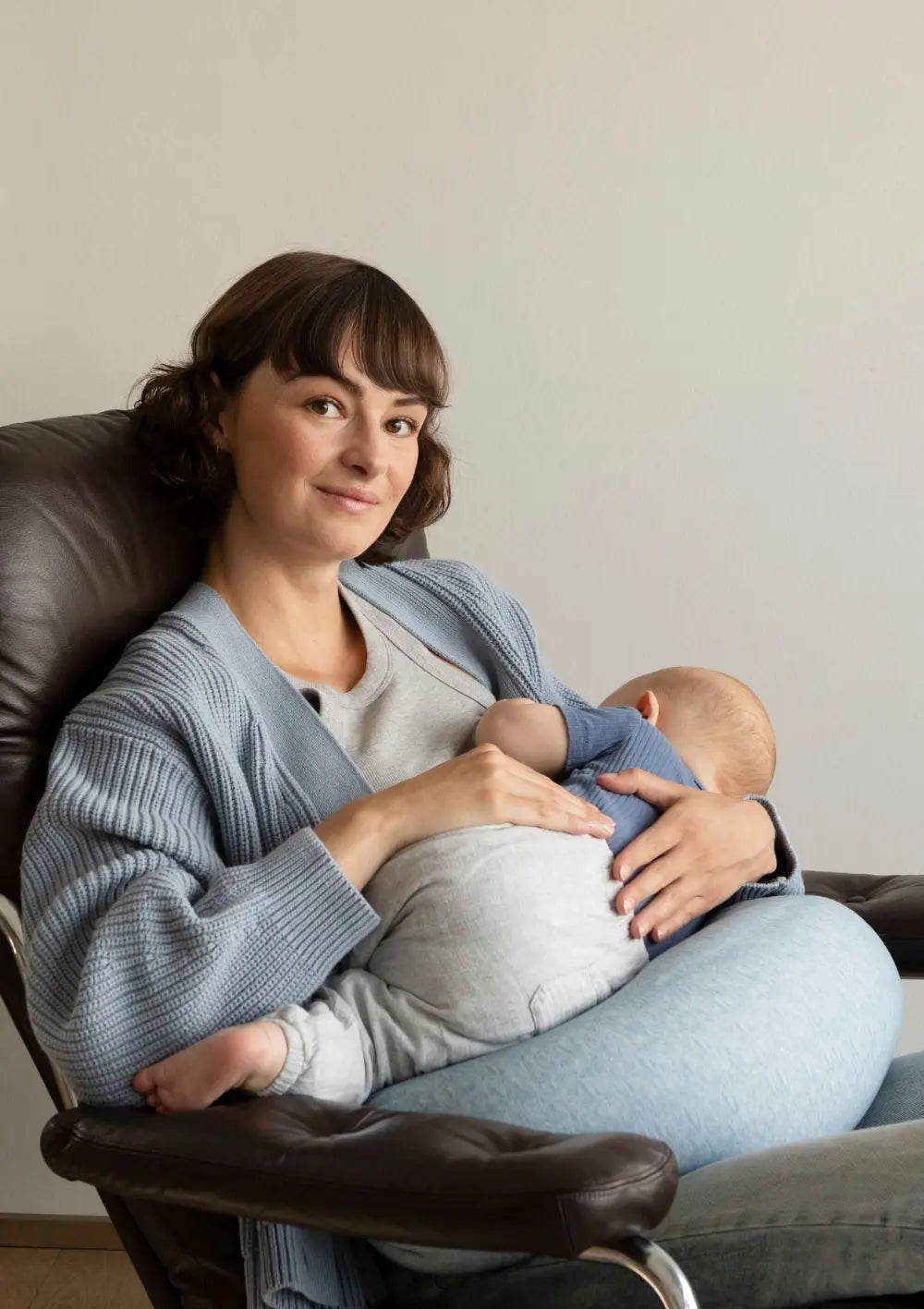 Lächelnde Mutter sitzt in einem braunen Ledersessel und stillt ihr Baby mit dem bbhugme Stillkissen Blue Mélange als Unterstützung.