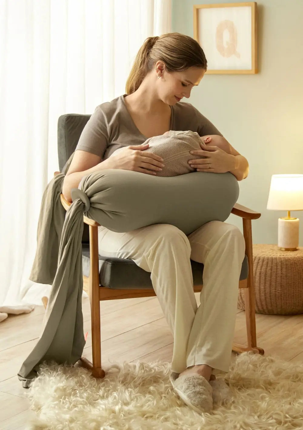 Eine Frau sitzt auf einem Stuhl mit ihrem Baby im Arm und wird gestützt vom bbhugme Schwangerschaftskissen Dusty Olive.
