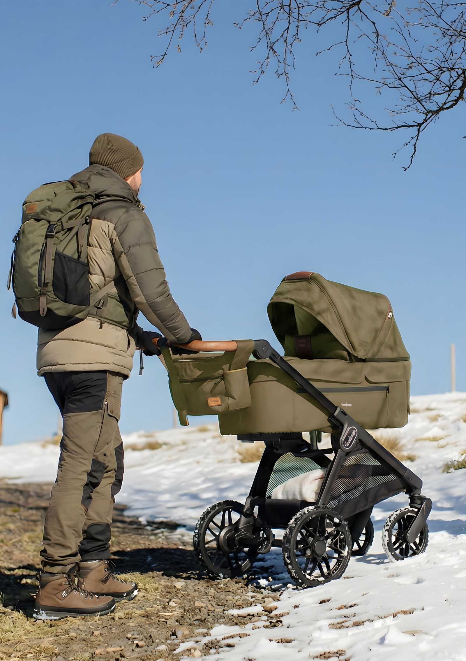 Mann schiebt Emmaljunga Kinderwagen 'SENTO PRO' Outdoor Olive in verschneiter Hügellandschaft