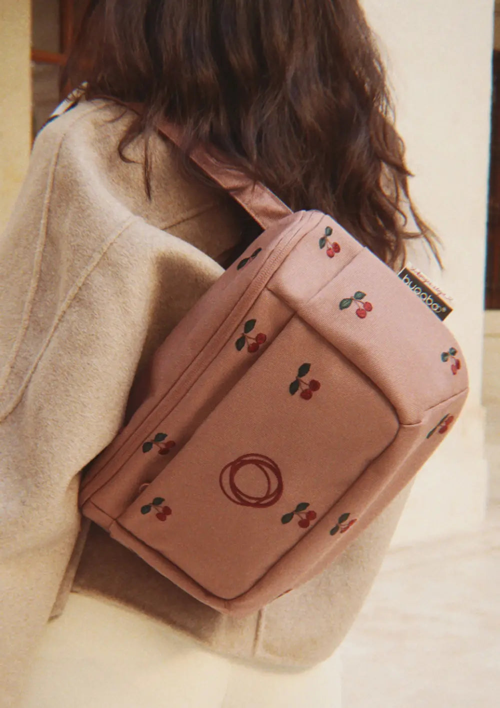 Detailansicht des Bugaboo Organizer x Konges Sløjd Dusty Pink Cherry, getragen als kompakte Umhängetasche über der Schulter; die Tasche zeigt eingestickte Kirschen auf altrosa Grund.
