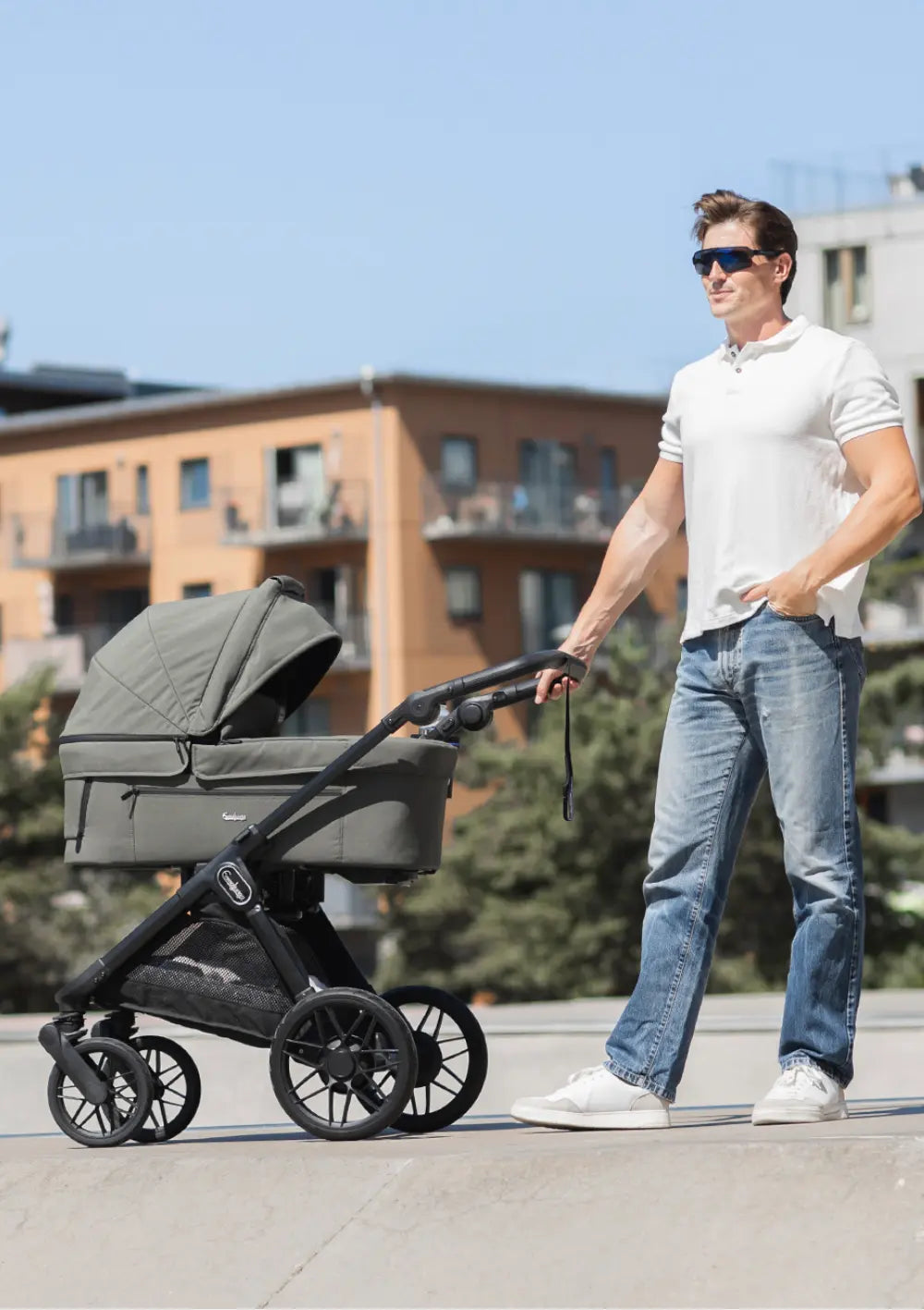 Ein Mann in Poloshirt und Sonnenbrille schiebt den olivenfarbenen Kombikinderwagen mit Babywanne Emmaljunga Kinderwagen SENTO PRO Urban Green über einen städtischen Platz.