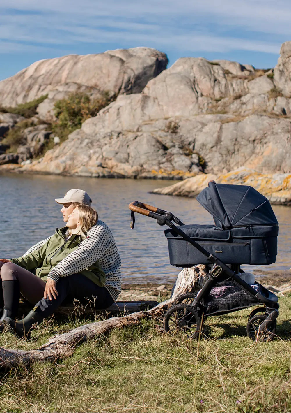 Ein Paar sitzt entspannt an einem felsigen Ufer im Freien, während der dunkelblaue Kombikinderwagen des Emmaljunga Kinderwagen-Set SENTO PRO Outdoor Navy neben ihnen steht.