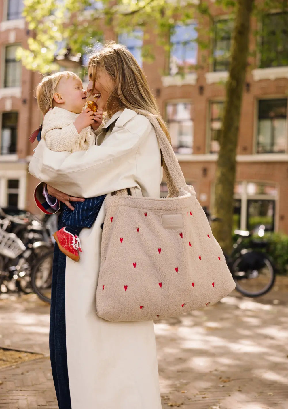 Eine Mutter trägt die beigefarbene Studio Noos Teddy-Shopper-Tasche mit Herzen-Muster über der Schulter und lacht mit ihrem Kind auf dem Arm im Freien.