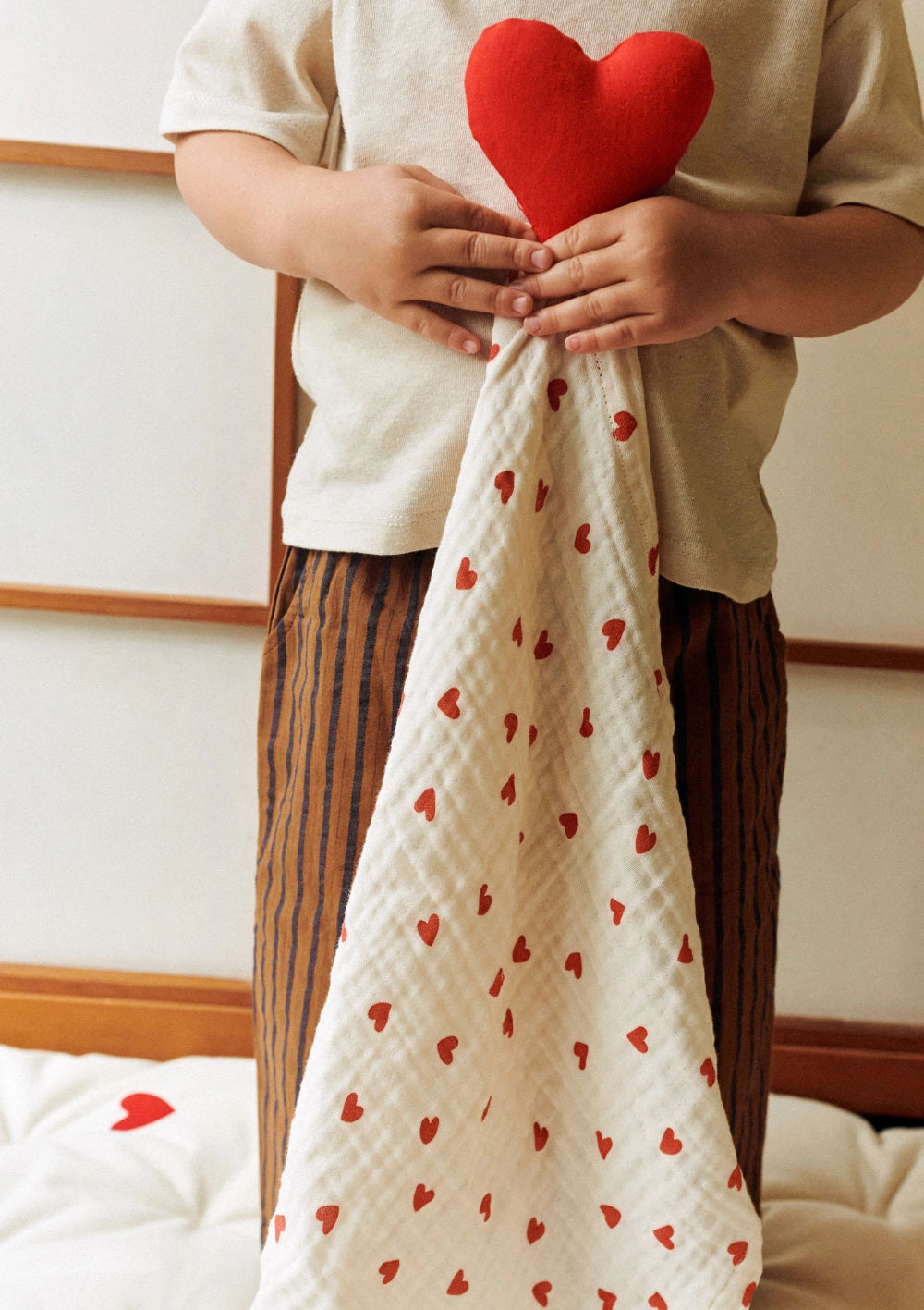 Ein Kind hält das Dear April Kuscheltuch Aura Little Love aus weißem Musselin mit roten Herzen und einem roten Stoffherz am Ende fest umschlungen.