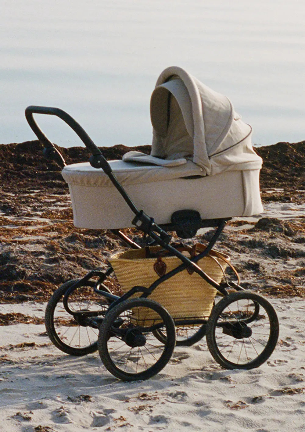 Angelcab Kinderwagen 'C3-Series' Upminster steht auf Sand, dahinter sind Felsen. Der Korb ist mit einer großen Korbtasche beladen.