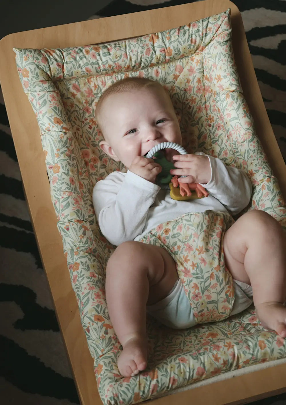 Baby mit Spielzeug liegt in der Charlie Crane Babywippe 'LEVO' Mushie Pastel Bloom (Buche)
