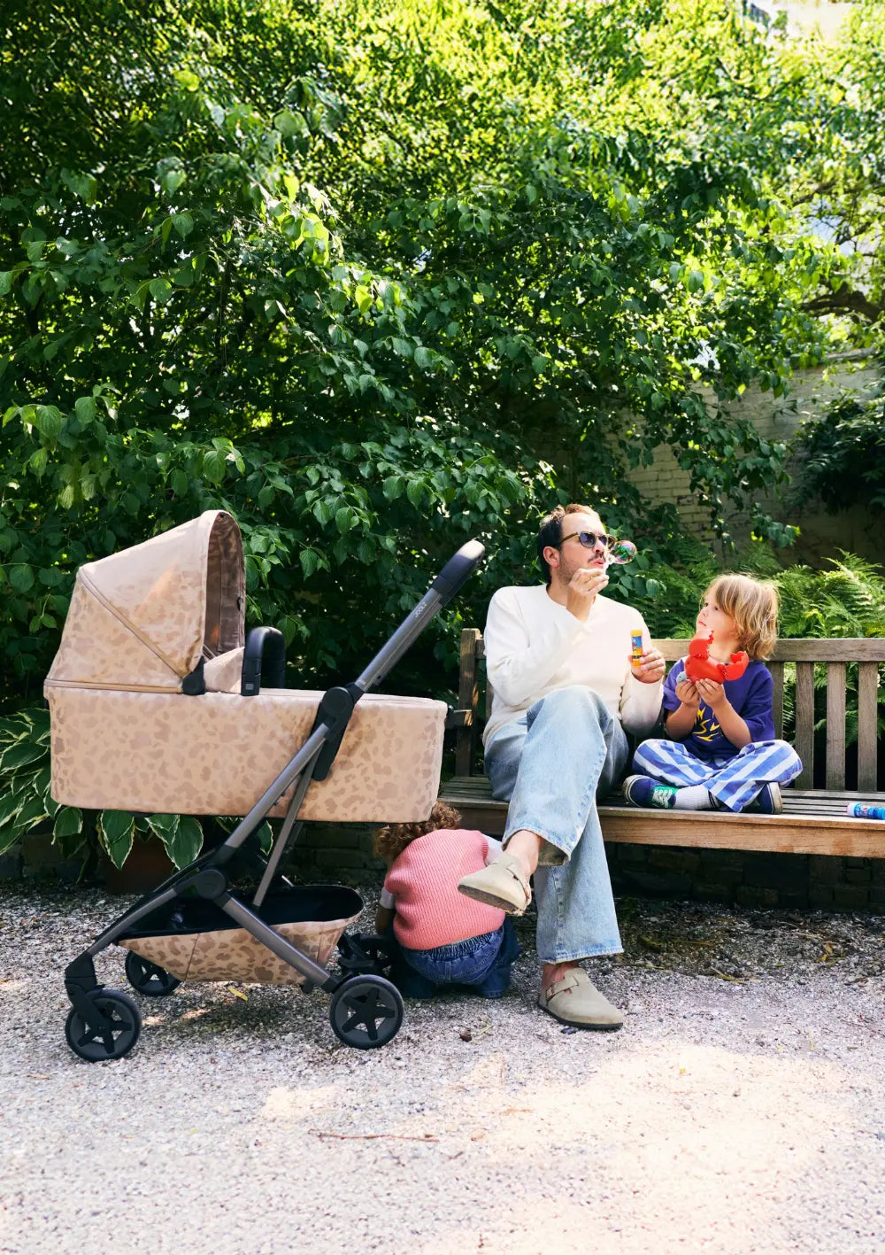 Ein Vater bläst auf einer Parkbank Seifenblasen für seine Kinder, während der beigefarbene Joolz Kinderwagen Aer2 Chic Renaissance Sandy Taupe mit passender Babywanne daneben steht.