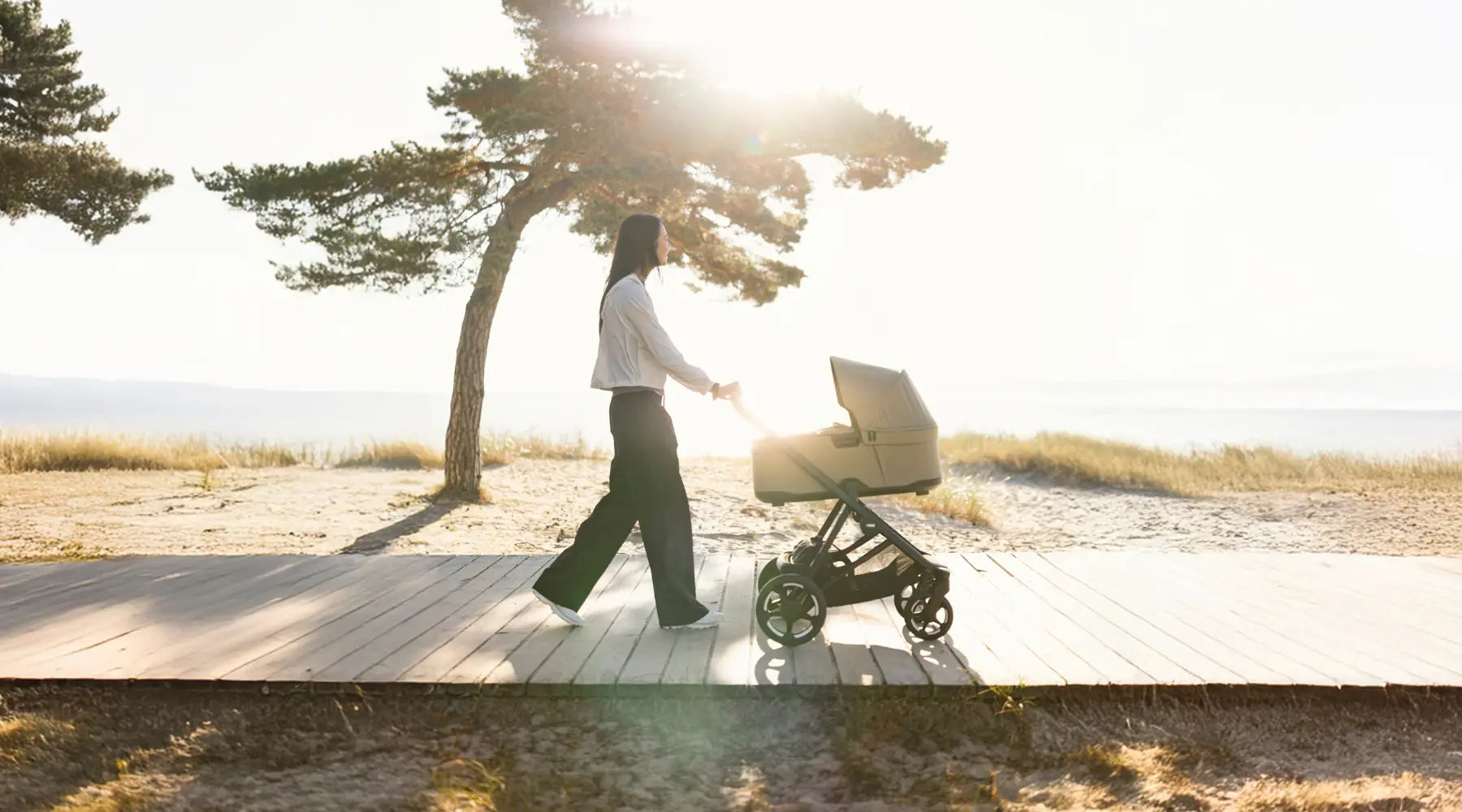 Eine junge Frau schiebt das Kinderwagen-Set 'Charm' in Faded Khaki über einen Holzsteg am Strand, während im Hintergrund eine Kiefer und das Meer im sanften, dunstigen Licht der tiefstehenden Sonne zu sehen sind.