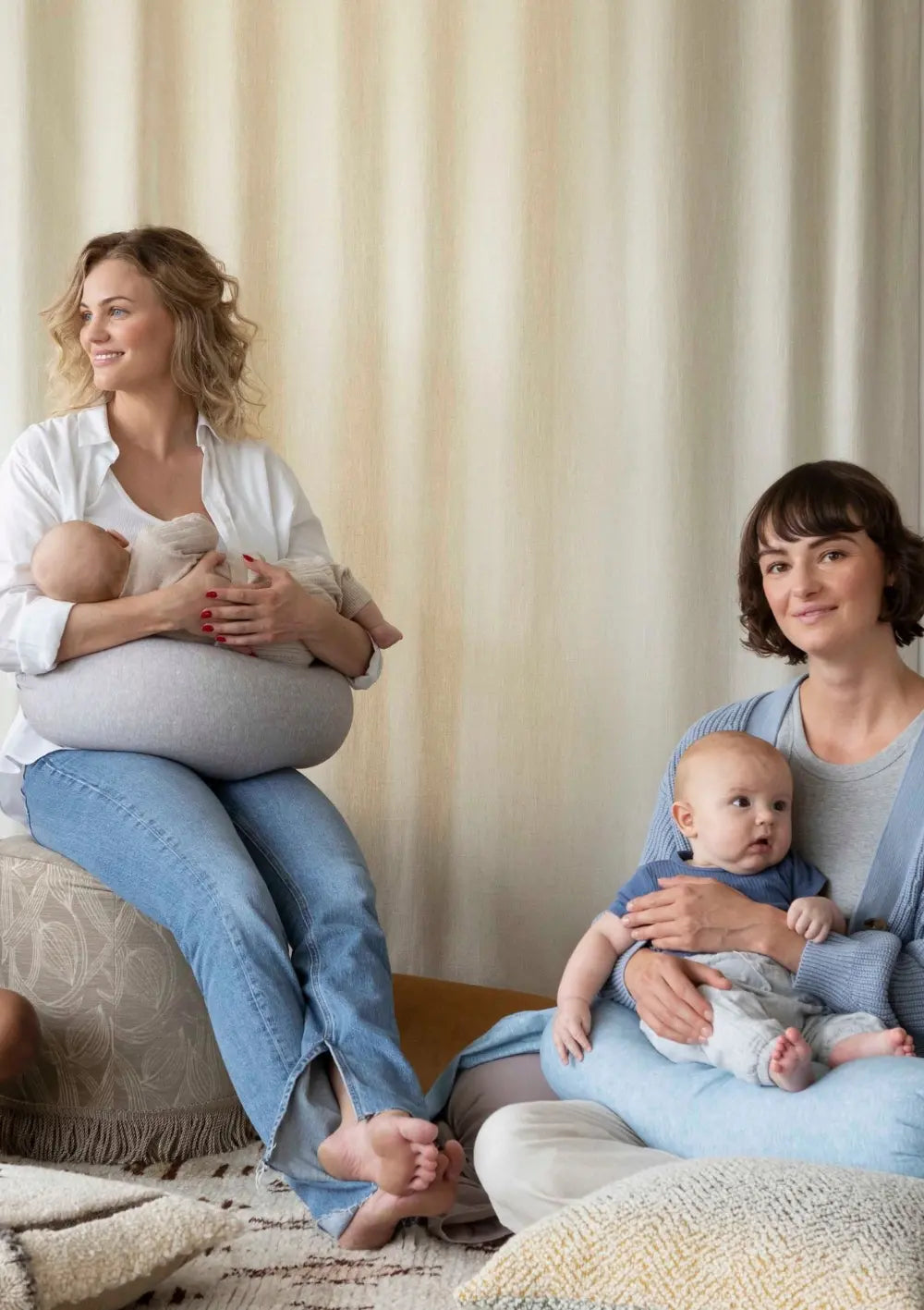 Zwei Mütter sitzen zusammen in einem hellen Raum mit ihren Babys – eine stillt mit dem bbhugme Stillkissen als Unterstützung, die andere hält ihr Baby auf dem Schoß.