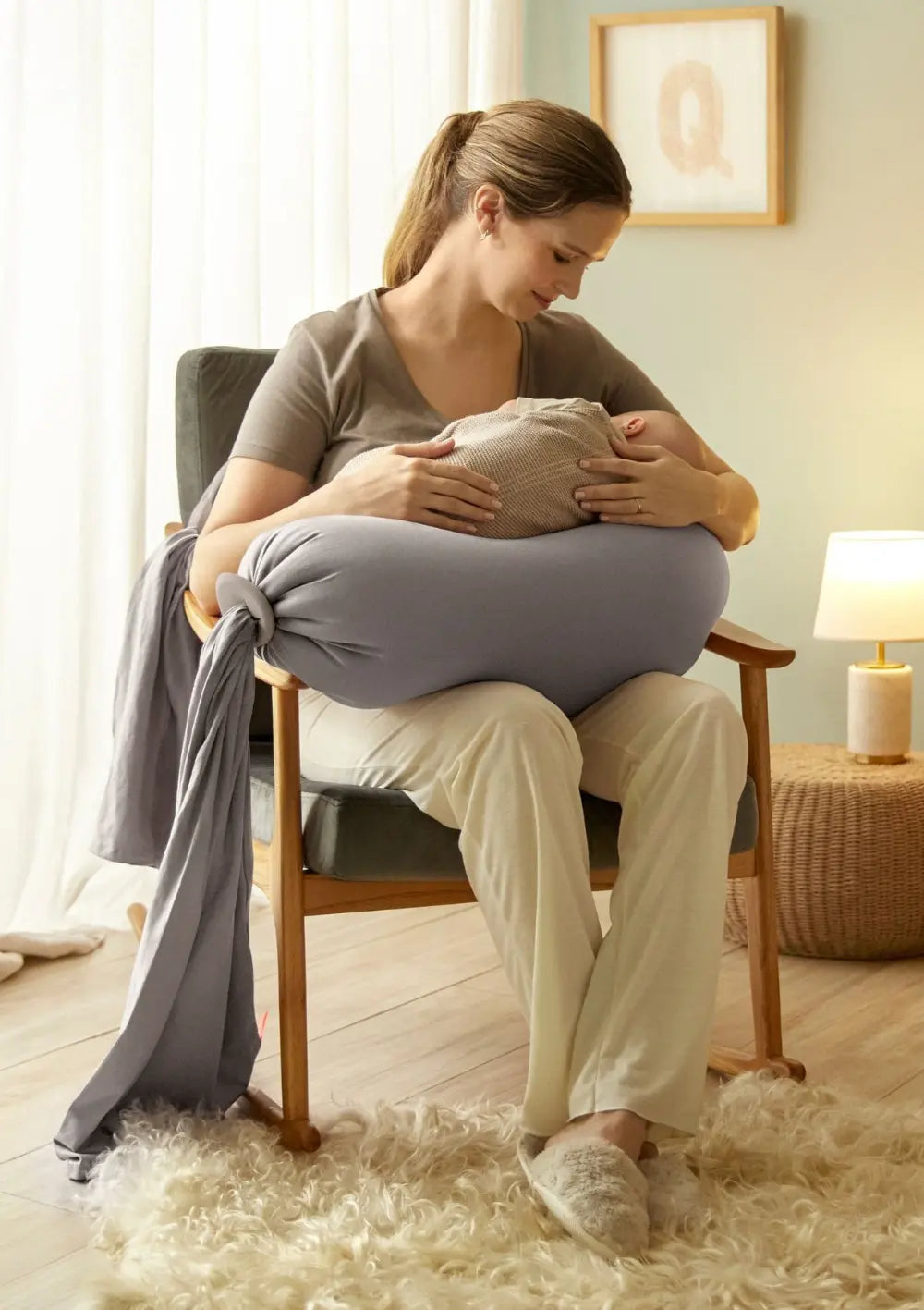 Eine Frau sitzt auf einem Stuhl mit ihrem Baby im Arm und wird gestützt vom bbhugme Schwangerschaftskissen Stone