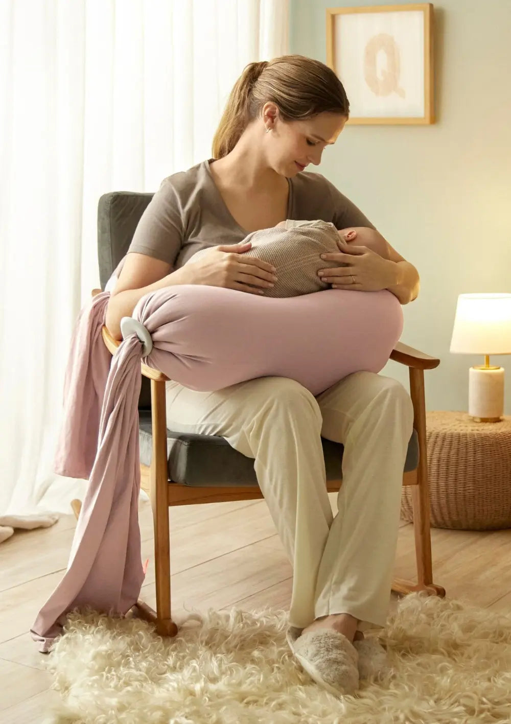 Eine Frau sitzt auf einem Stuhl mit ihrem Baby im Arm und wird gestützt vom bbhugme Schwangerschaftskissen Dusty Pink