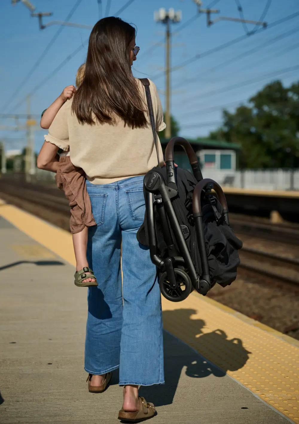 Rückenansicht einer Frau auf einem Bahnhofssteig, die ein Kleinkind auf der Hüfte trägt. Über der Schulter trägt sie den kompakt zusammengeklappten Nuna VIAA cabn Buggy mit dem markanten braunen Kunstledergriff. Die Szene betont die Eignung des Buggys für den unkomplizierten Einsatz im öffentlichen Nah- und Fernverkehr.