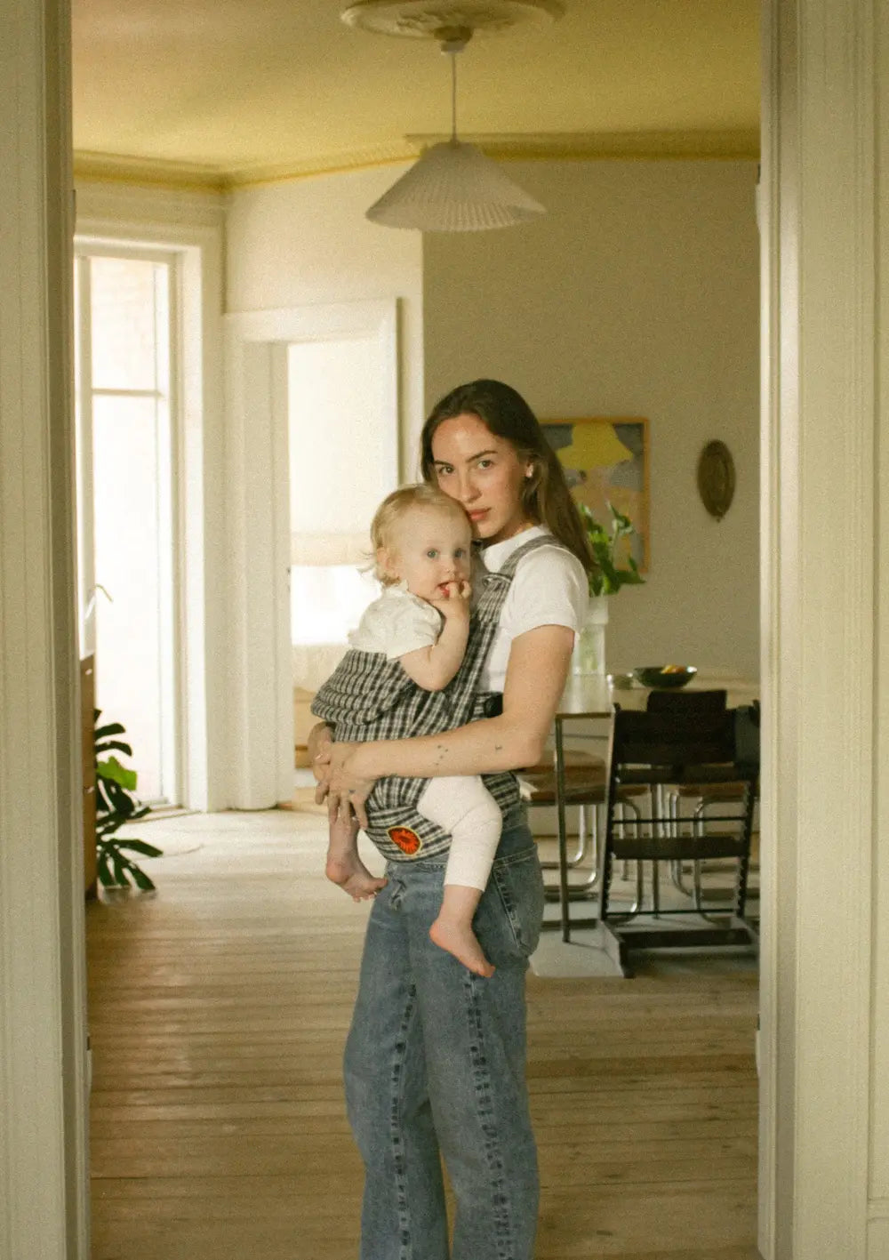 Eine Mutter im Wohn-/Essbereich einer Altbauwohnung trägt ihr Kind in der Moova Babytrage 'Balance' Navy Check