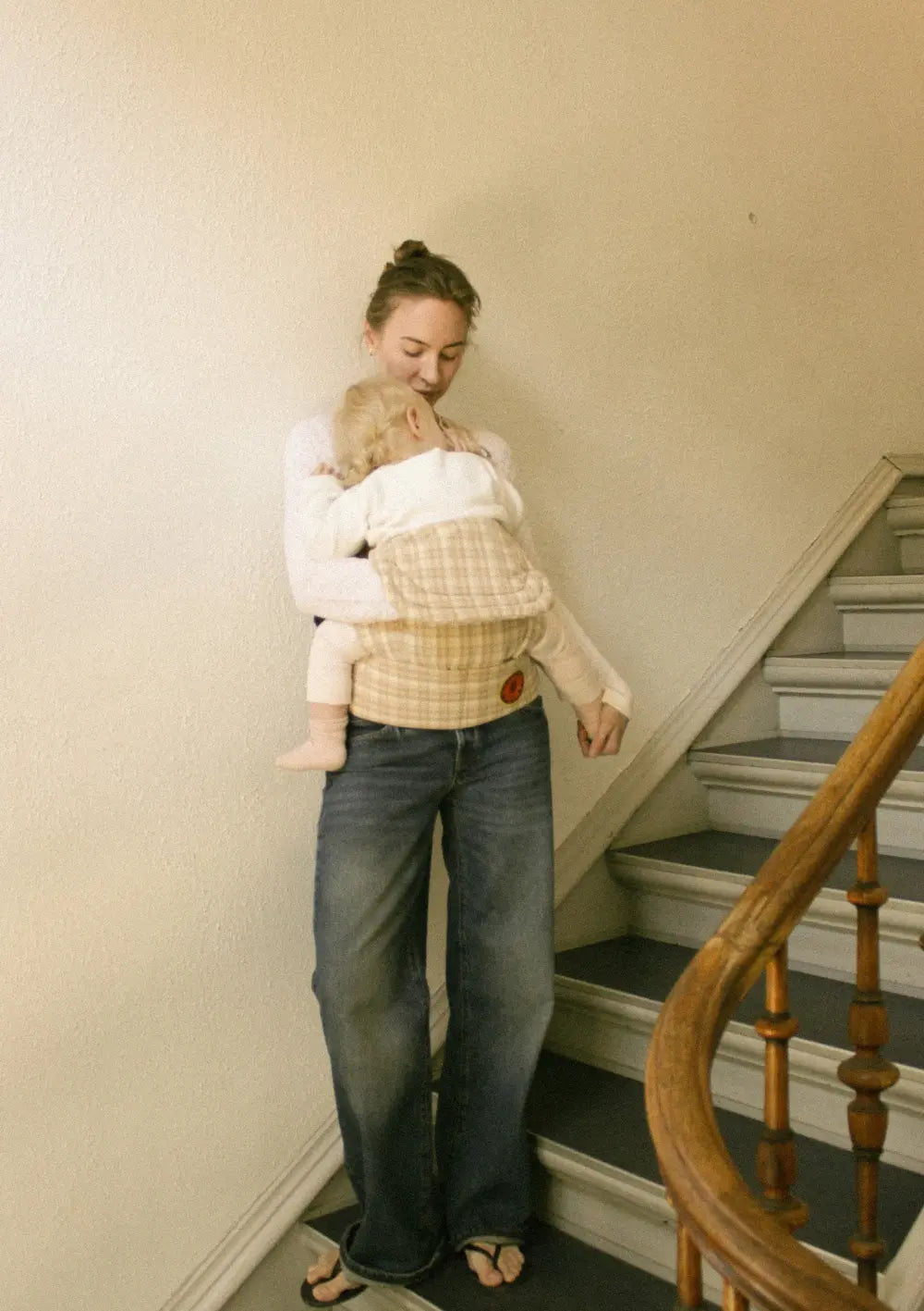 Eine Mutter steht in einem Treppenhaus und trägt ihr schlafendes Kind in der Moova Babytrage 'Balance' Mocha Check