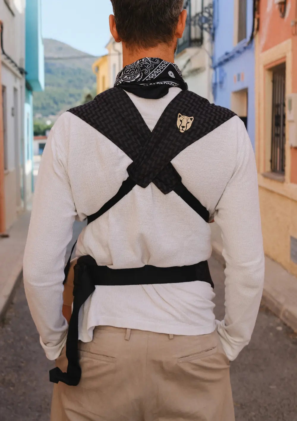 Rückansicht eines Mannes in einer Gasse, der die gekreuzten Träger und den Hüftgurt der Wildride Babytrage Firstride Bowie Minimal Schwarz mit dezentem Webmuster trägt.