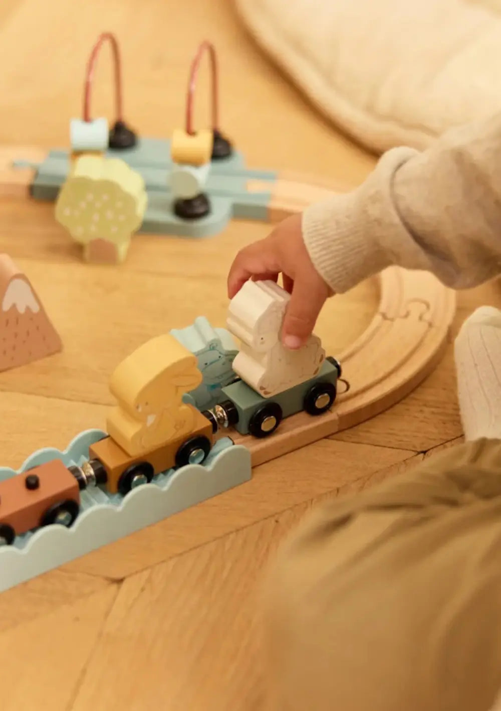 Ein Kind setzt ein hölzernes Schaf auf einen Zugwaggon der Jollein Holz Eisenbahn Set Jollies, die auf Holzschienen auf dem Boden aufgebaut ist.